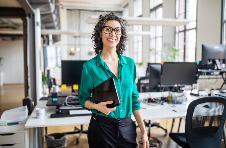 A woman smiling while holding a tablet, at her back there are computer desktops and other office equipments